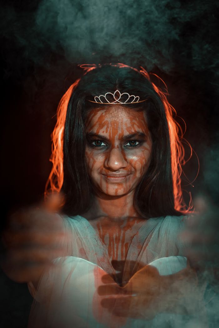 A woman with eerie makeup and tiara, casting an unsettling smile in a dark, smoke-filled setting.