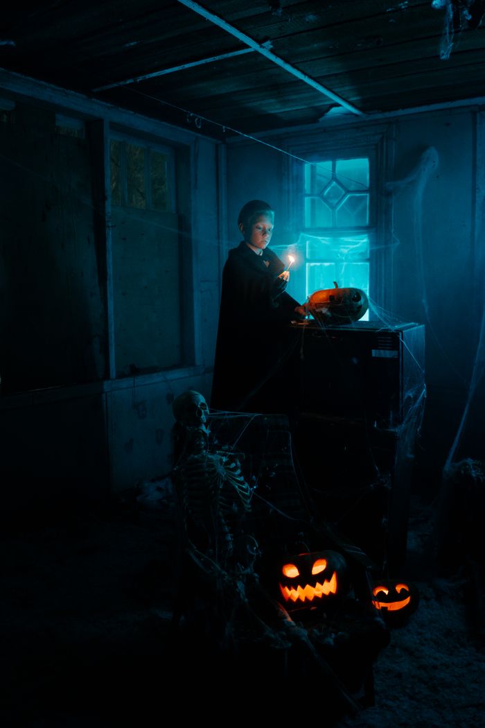 A boy in a dimly lit room with Halloween pumpkins and a skeleton, creating a spooky atmosphere.